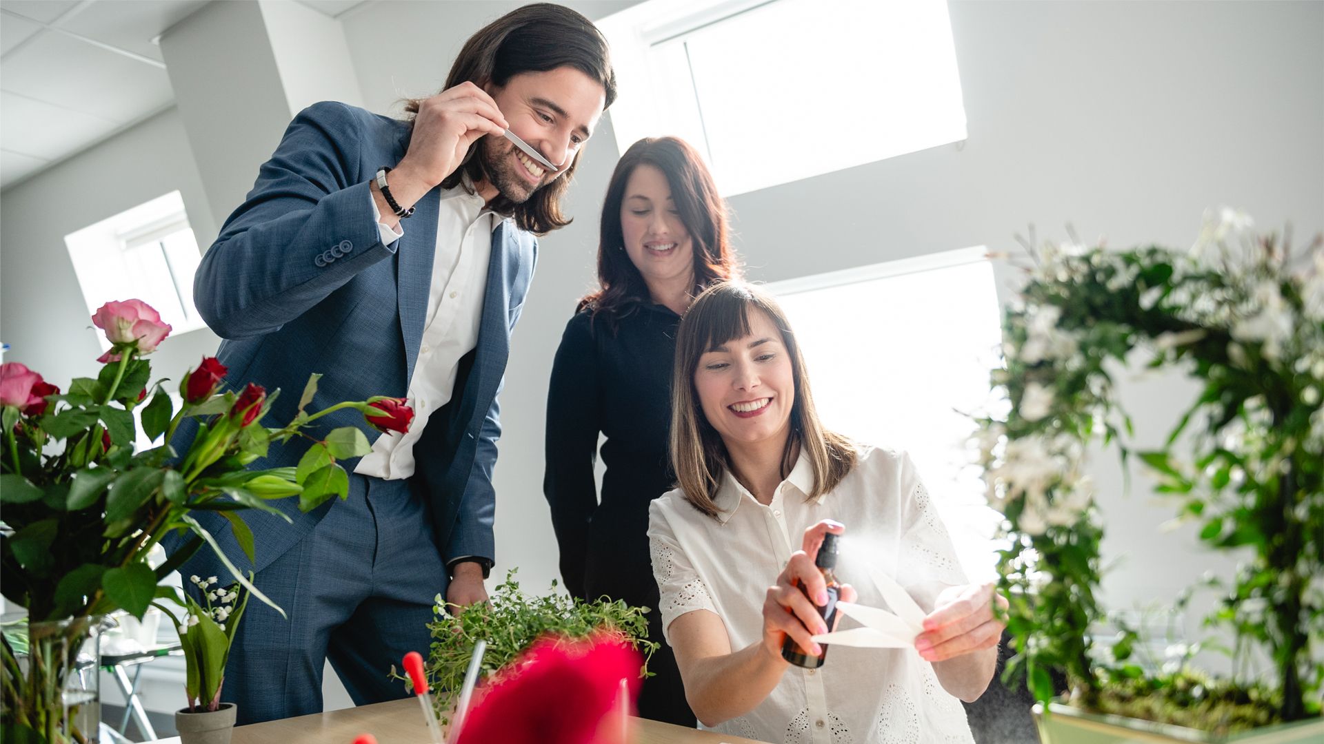 Three people working together at a table surrounded by flowers and plants. One person in a blue suit is holding a strip of card to their nose, another is partially visible in the middle, and a third person in a white blouse is spraying fragrance onto several strips of card arranged in a fan in their hand. The setting is well-lit with large windows in the background.