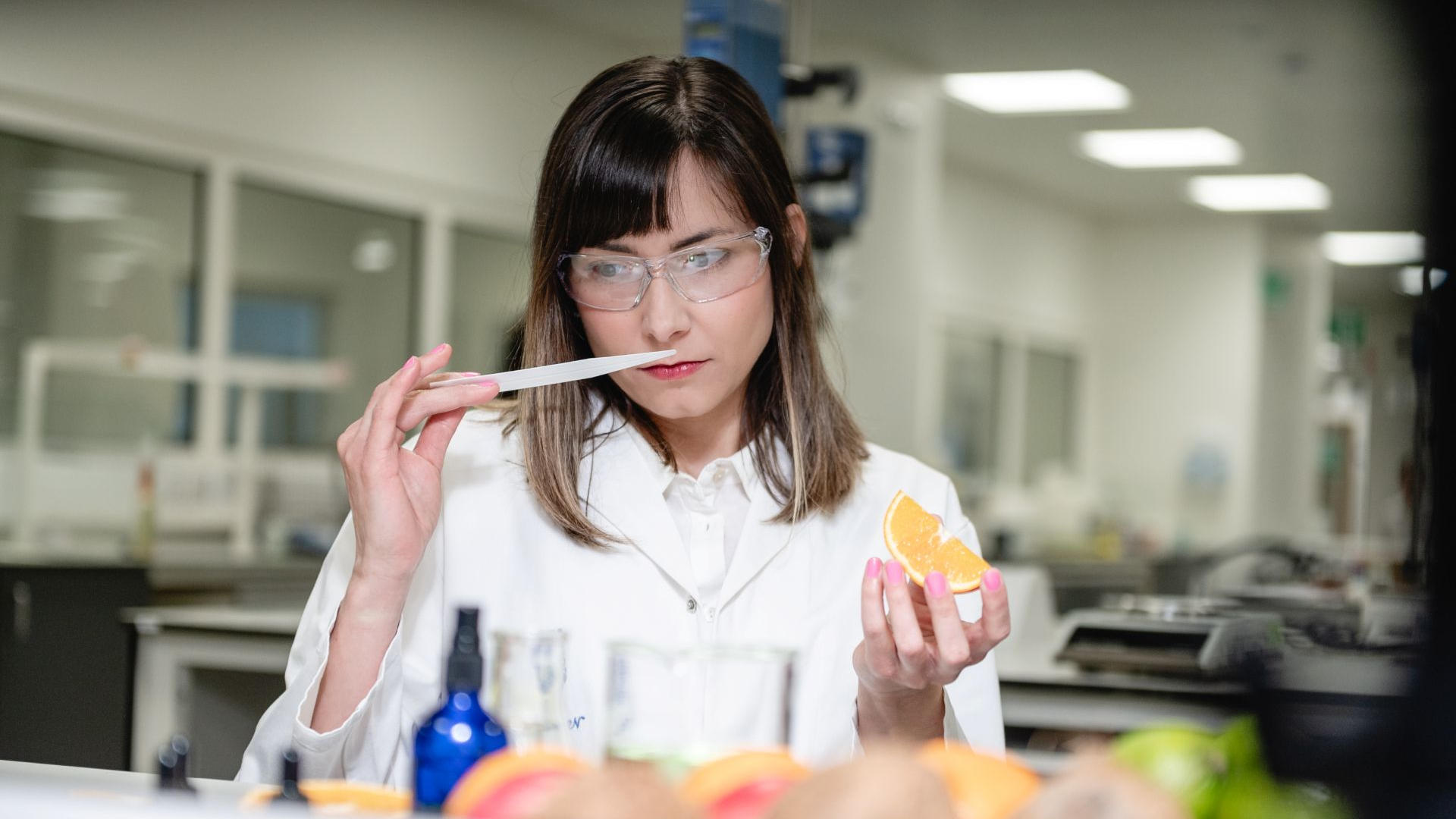 A woman in a lab coat and safety googles smells a paper-smelling strip while holding an orange  in her other hand.