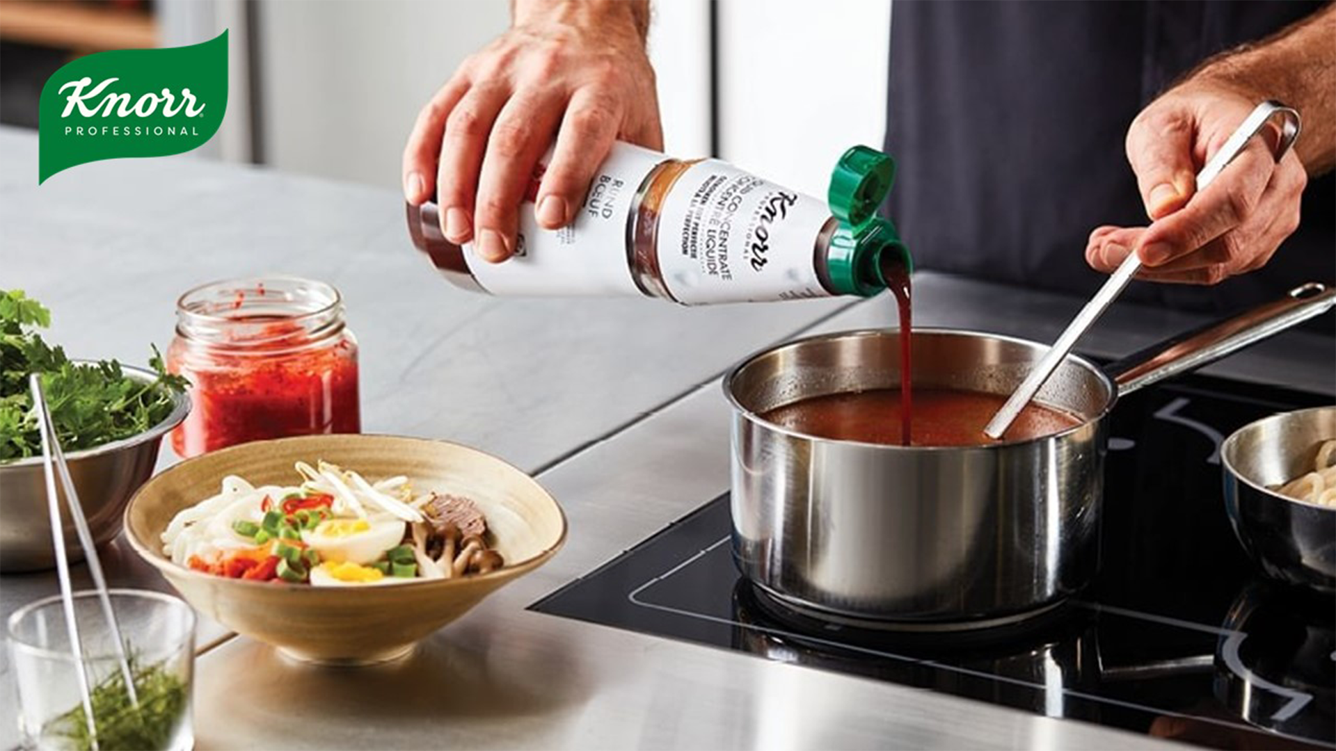 A person is cooking using an induction hob. They are stirring a pan with one hand while pouring a bottle of Knorr-branded product into the pot. Additional bowls of ingredients are arranged on the worktop.