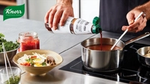 A person is cooking using an induction hob. They are stirring a pan with one hand while pouring a bottle of Knorr-branded product into the pot. Additional bowls of ingredients are arranged on the worktop.
