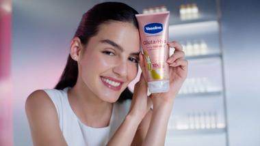 A white woman with dark hair and glowing skin, holding a tube of Vaseline Gluta Hya Dewy Radiance cream. Pink, premium packaging.