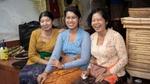 Three women in traditional clothing sit on a tiled step. They are surrounded by baskets, bamboo sticks, and household items in an outdoor setting.