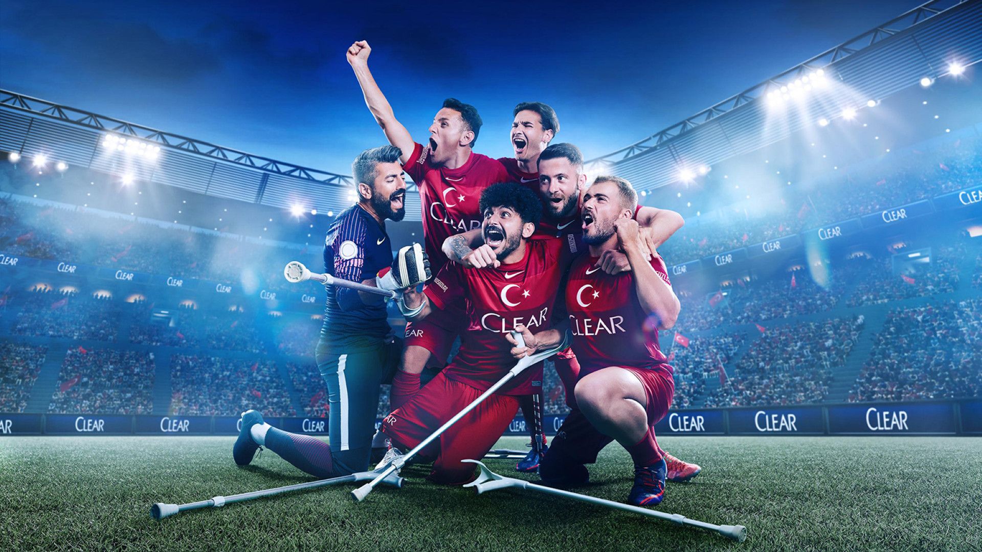 A group of six athletes on a grass sports field. Five are wearing a red kit; the sixth is wearing a purple shirt and dark green shorts. Several crutches are in view, either being held or lying on the grass. The scene is set in a brightly lit stadium filled with cheering spectators, with the Clear brand logo displayed prominently in the background.