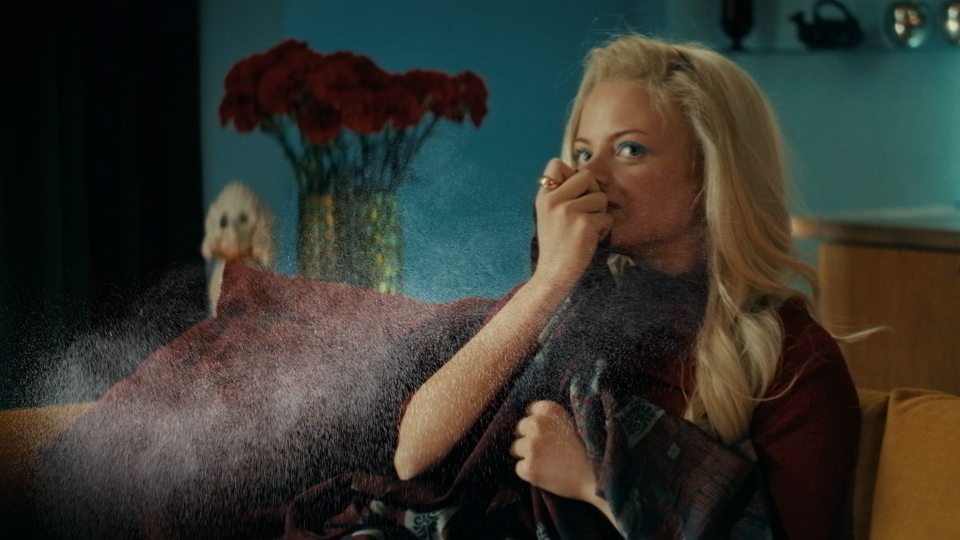  A person seated on a sofa holds a blanket close while leaning forward to smell a cloud of fine mist or spray that is dispersing in the air. A vase filled with red flowers is visible in the background against a blue wall.