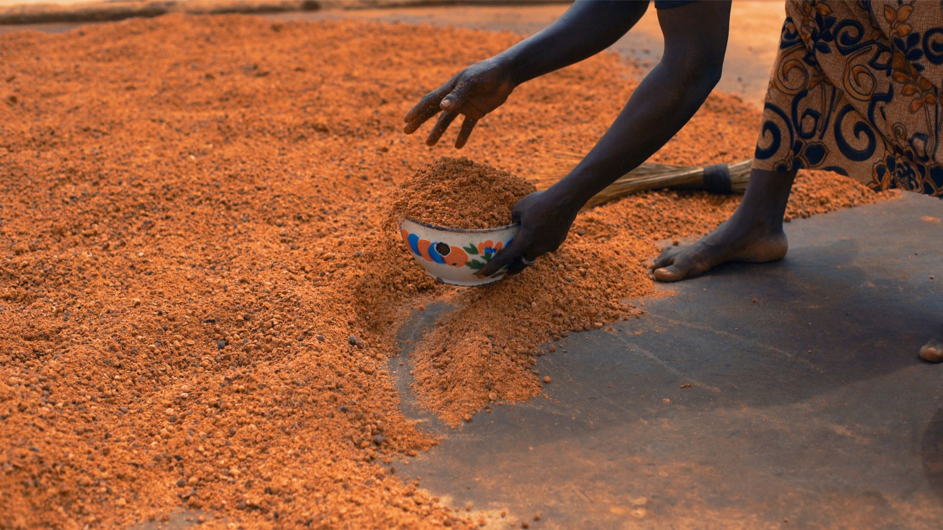 A woman making shea butter in Ghana. Her bare feet, hands and part of her pattered dress are visible. Ground shea nuts are in a bowl in her left hand.