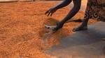 A woman making shea butter in Ghana. Her bare feet, hands and part of her pattered dress are visible. Ground shea nuts are in a bowl in her left hand.