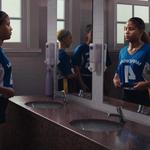 A group of girls in sports uniforms stand in a locker-room–style bathroom with sinks and mirrors. One girl adjusts her uniform while others stand nearby, reflected in the mirrors.