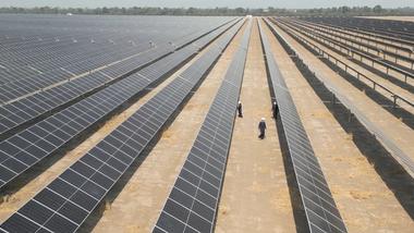 Rows of solar panels stretch into the distance. Three workers in safety gear and helmets walk between the rows, inspecting the panels.
