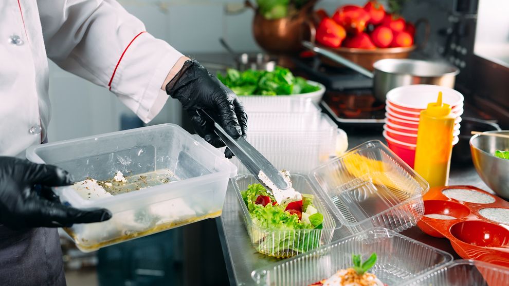 Chef in a commercial kitchen placing food into a takeaway container, with various bowls of ingredients on the work surface