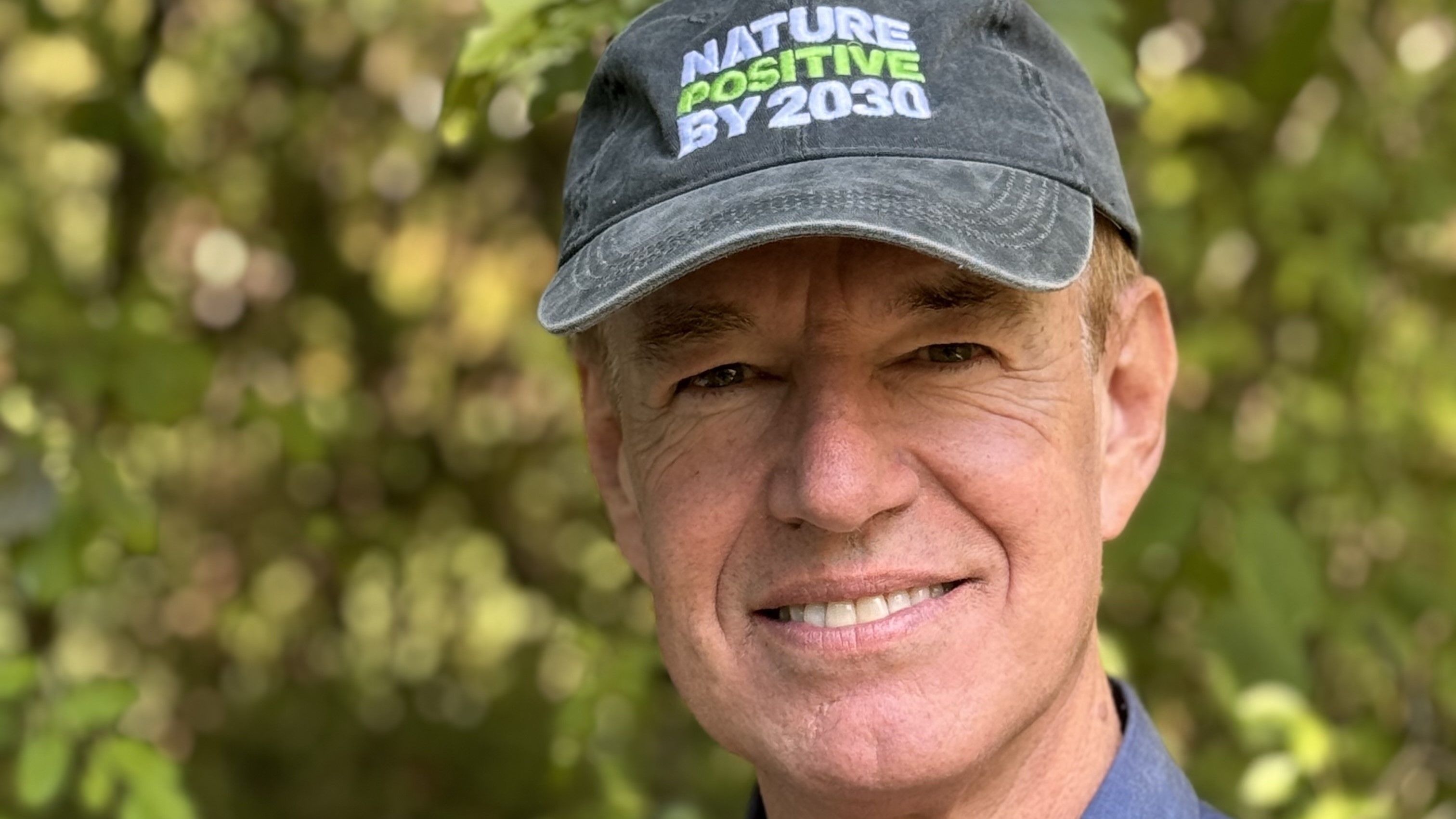 A headshot of a Marco Lambertini. Marco is stood outside with trees behind him. He is wearing a grey cap that says “Nature Positive by 2030” and a blue shirt. He is smiling at the camera.