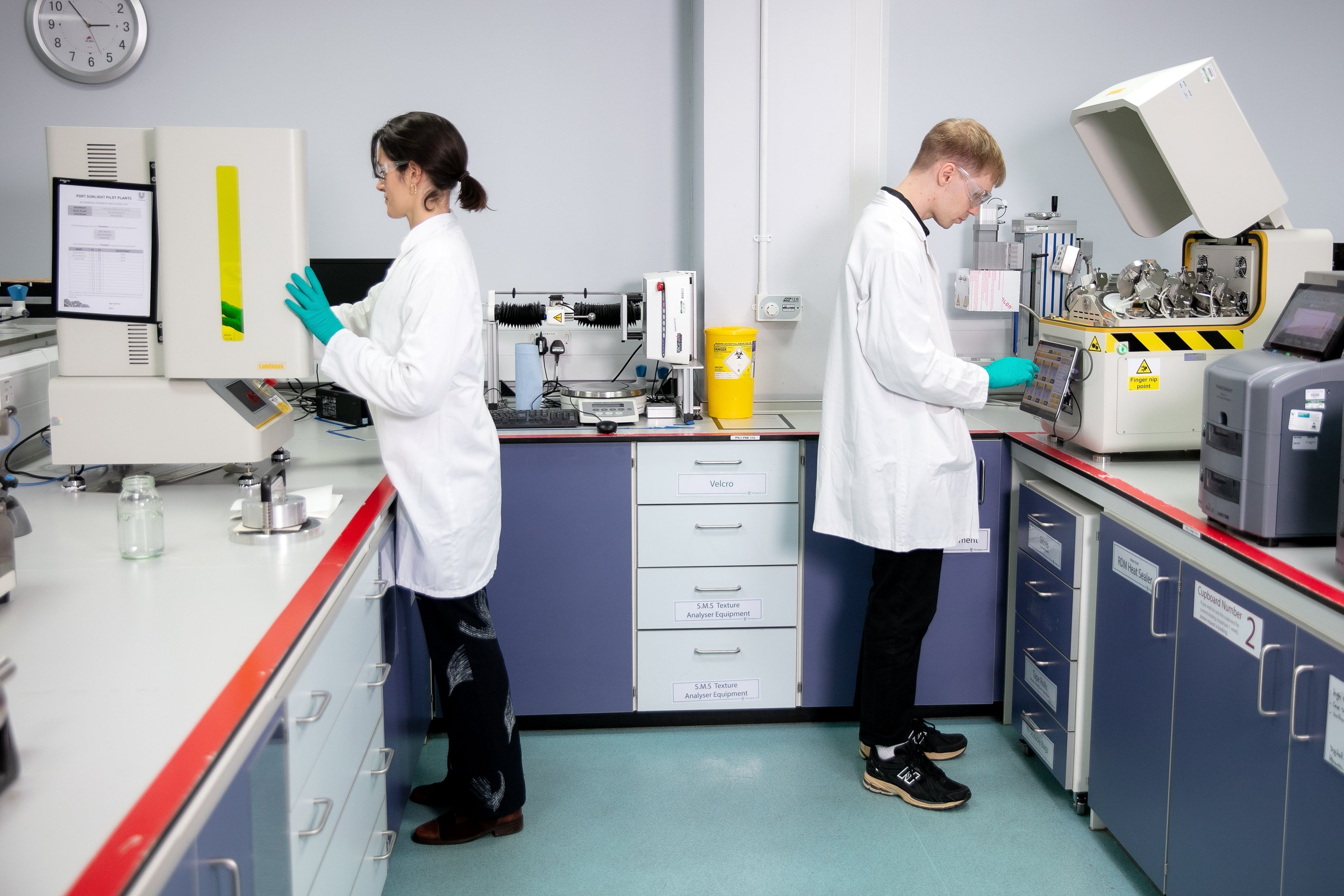 Two technicians in white lab coats and safety goggles working in a research lab, equipped with various devices, labelled drawers and containers.