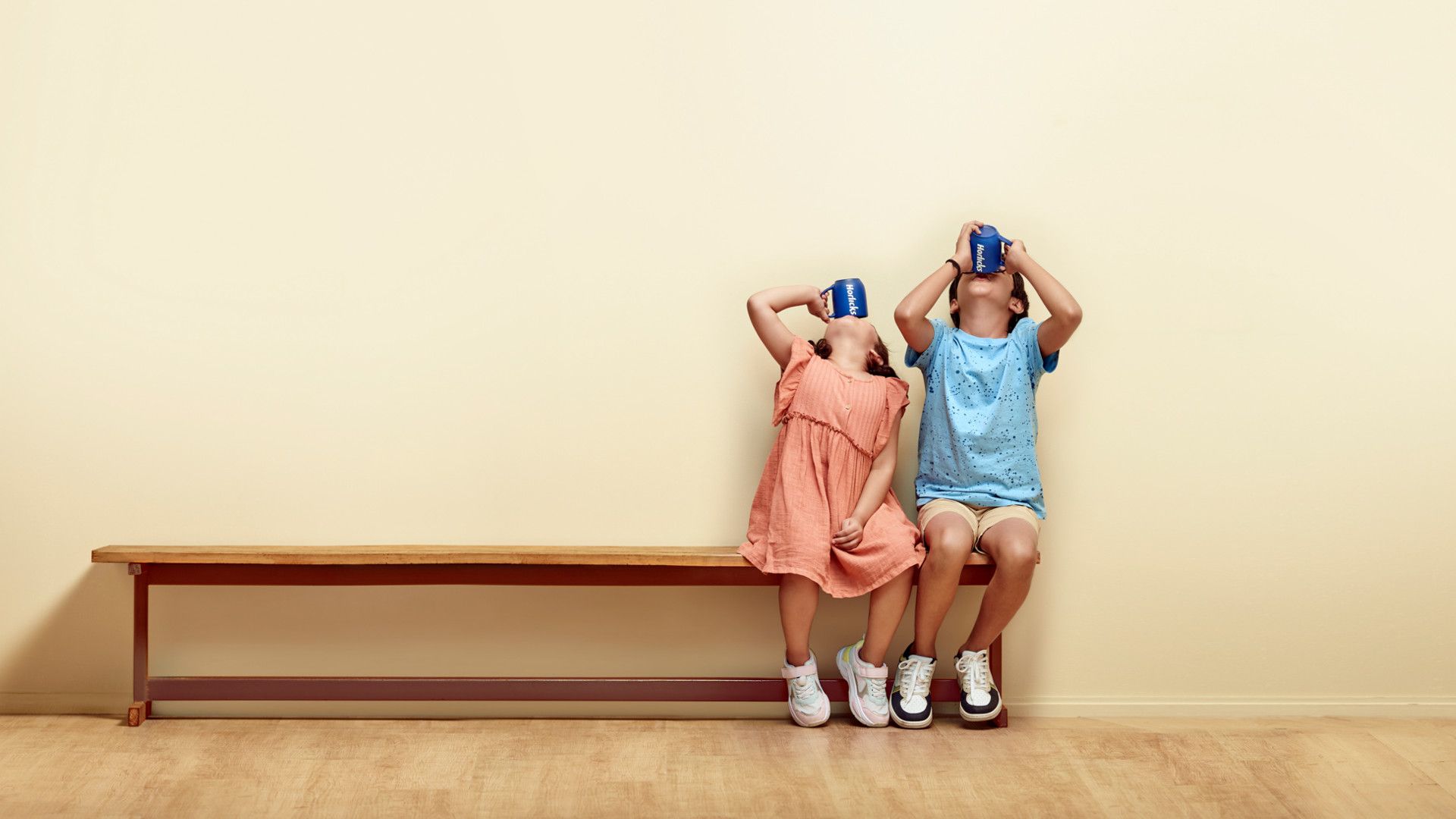 Boy and girl sitting on a bench with their heads thrown back to get every last drop of Horlicks from their cup