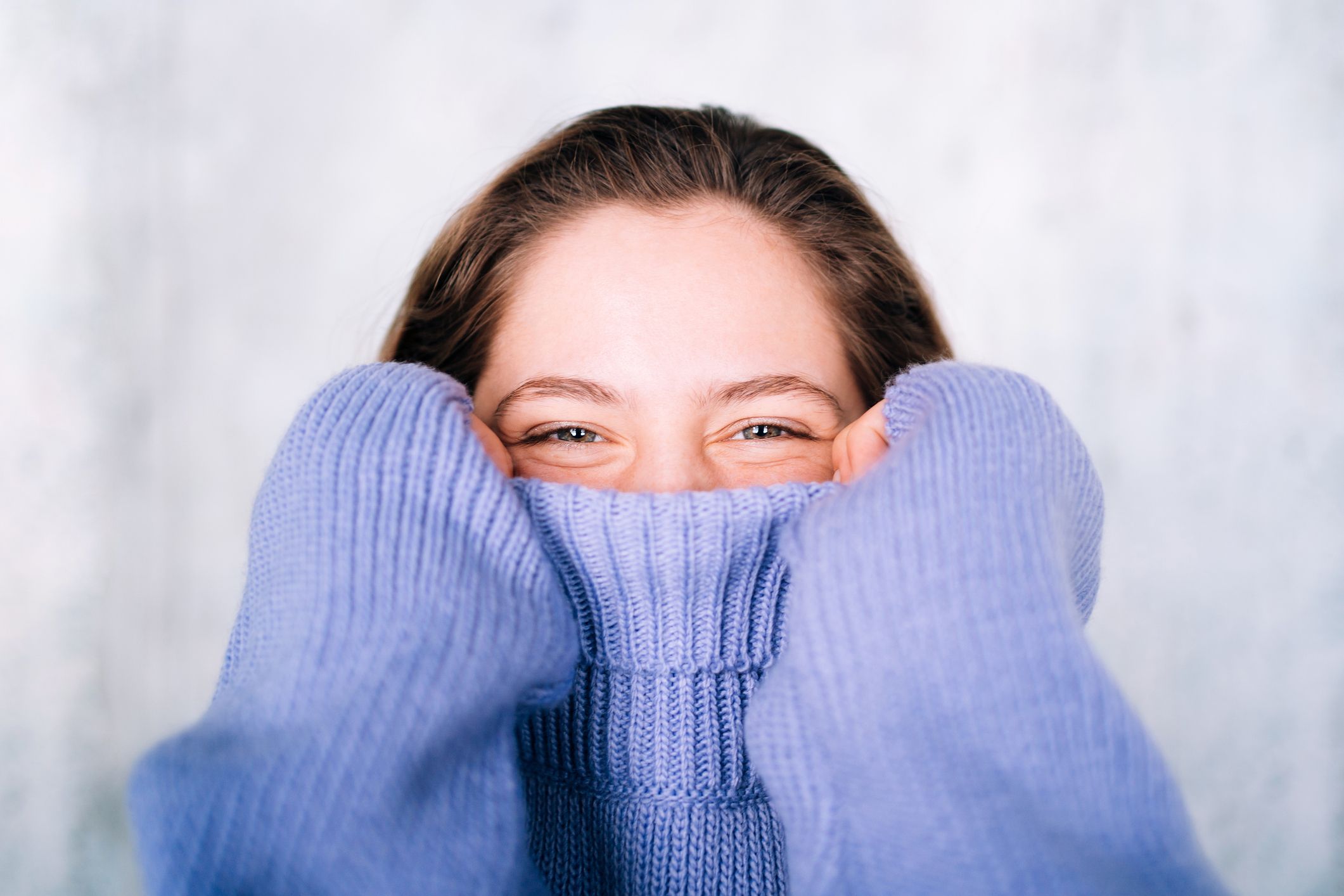 A woman pulls her cosy sweater up over her face to below her glinting eyes.