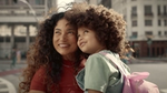 A woman holding a child with a pink backpack, with a city street in the background. They are both smiling.