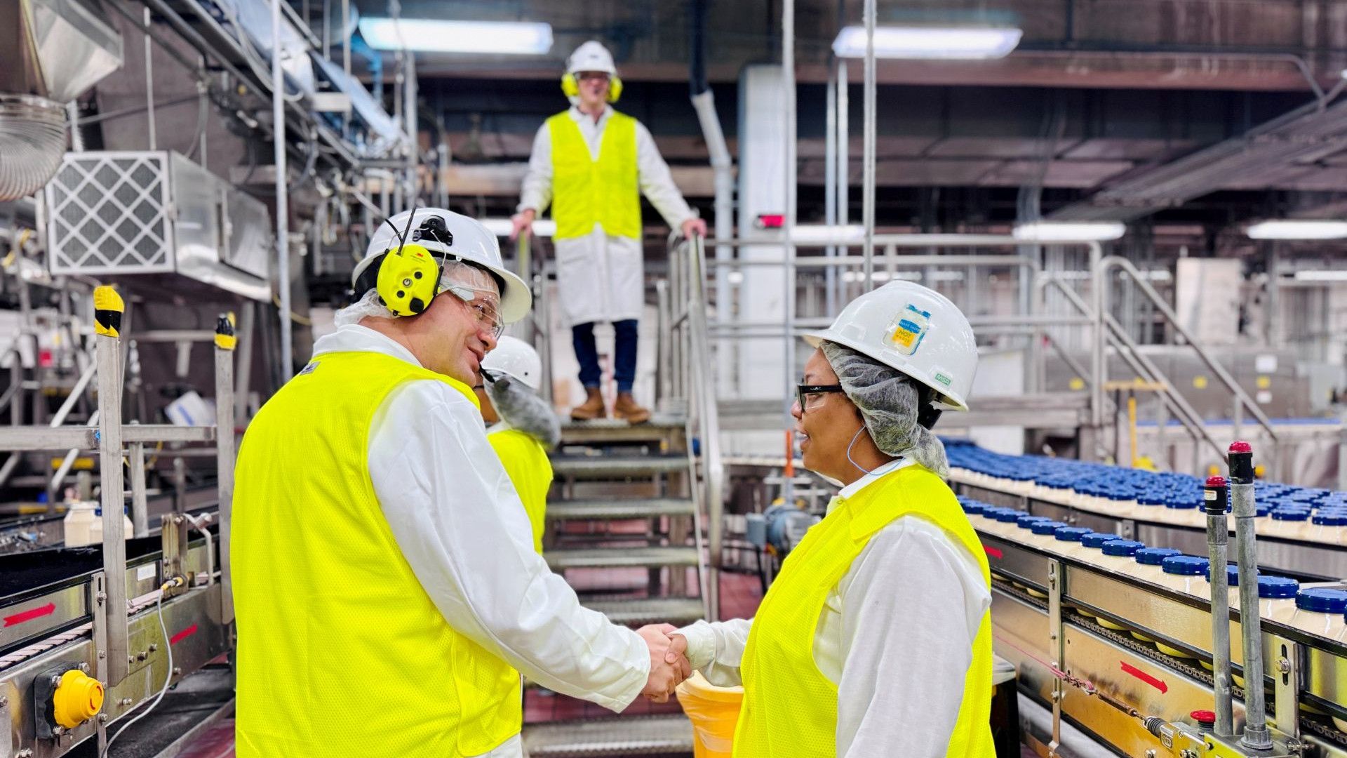 Two factory workers in yellow high-vis jackets and hard hats shake hands next to a Unilever factory production line.