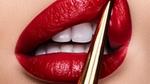Extreme close-up of glossy red lips with white teeth, applying lipstick using a gold applicator.