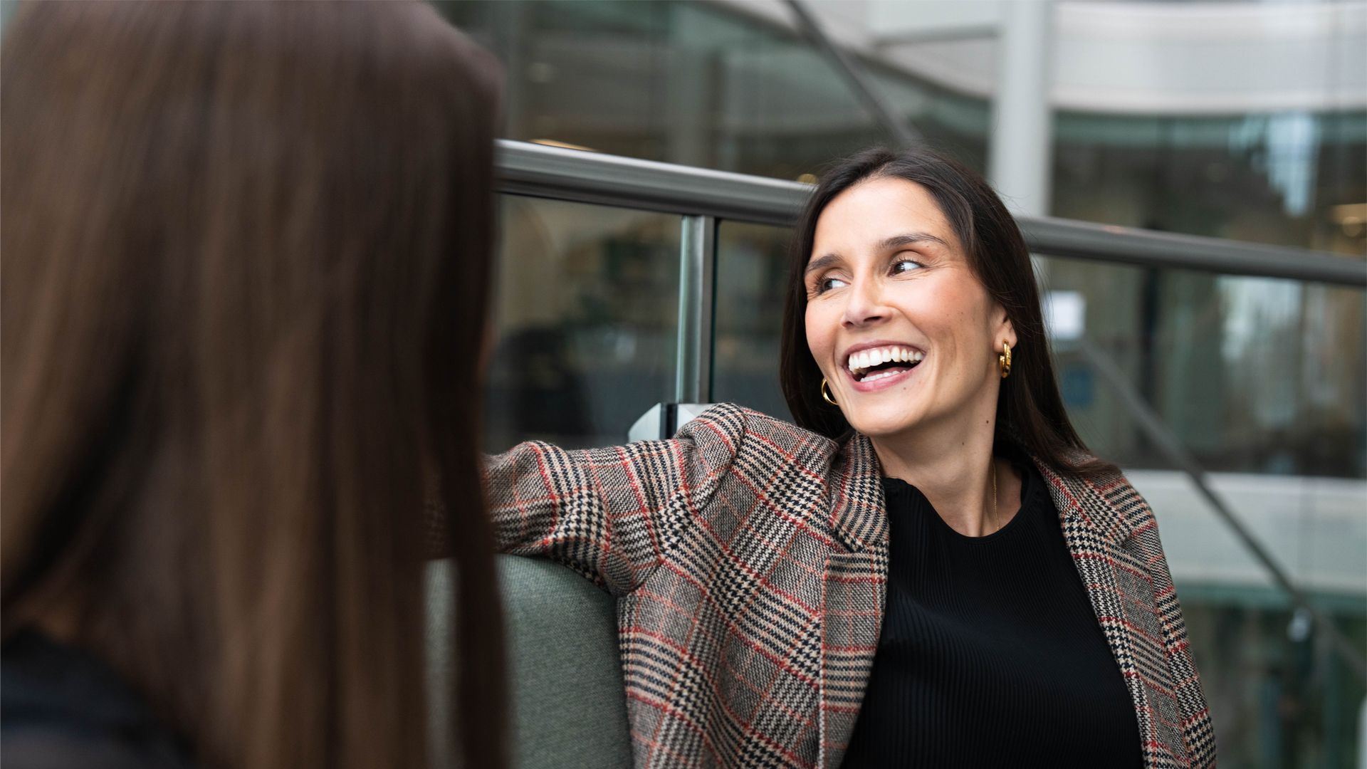 Two individuals are seated and conversing in a modern office space with glass railings and large windows. One person wears a plaid blazer over a black top and is smiling, while the other has long brown hair and is side on.