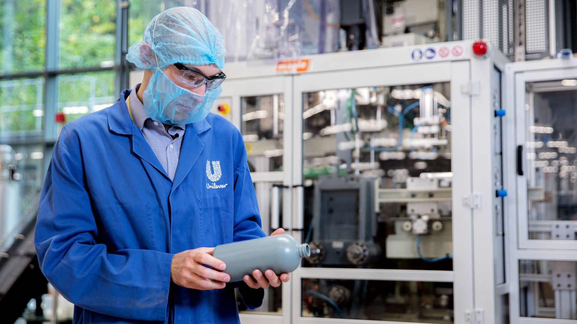 A person wearing a blue Unilever lab coat, hairnet, safety glasses and face mask examines a grey plastic bottle in a factory setting, with industrial equipment in the background.