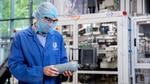 A person wearing a blue Unilever lab coat, hairnet, safety glasses and face mask examines a grey plastic bottle in a factory setting, with industrial equipment in the background.