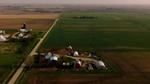 Aerial view of some rural farmland with several buildings including barns, surrounded by vast expanses of green and brown agricultural fields.