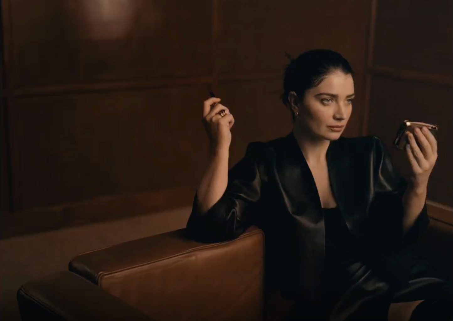 Woman sat on a brown couch dressed in black with a beauty product in her hand.