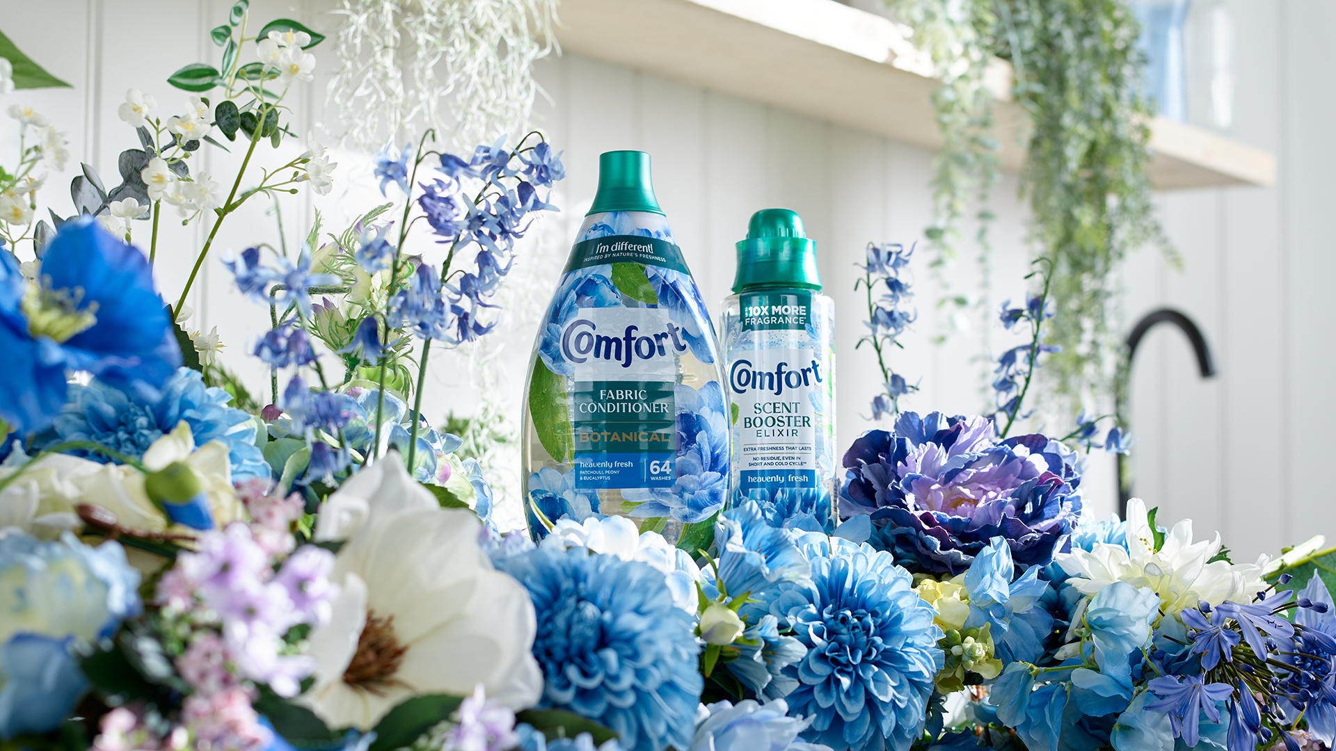 Two Comfort bottles from the Botanicals and Elixir ranges on a white shelf surrounded by blue, lavender, pink and white flowers.