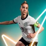 A football player wearing a white jersey with black trim and a green captain’s armband, posed in action with a ball at their feet. The background is teal with glowing white neon lines forming dynamic shapes.