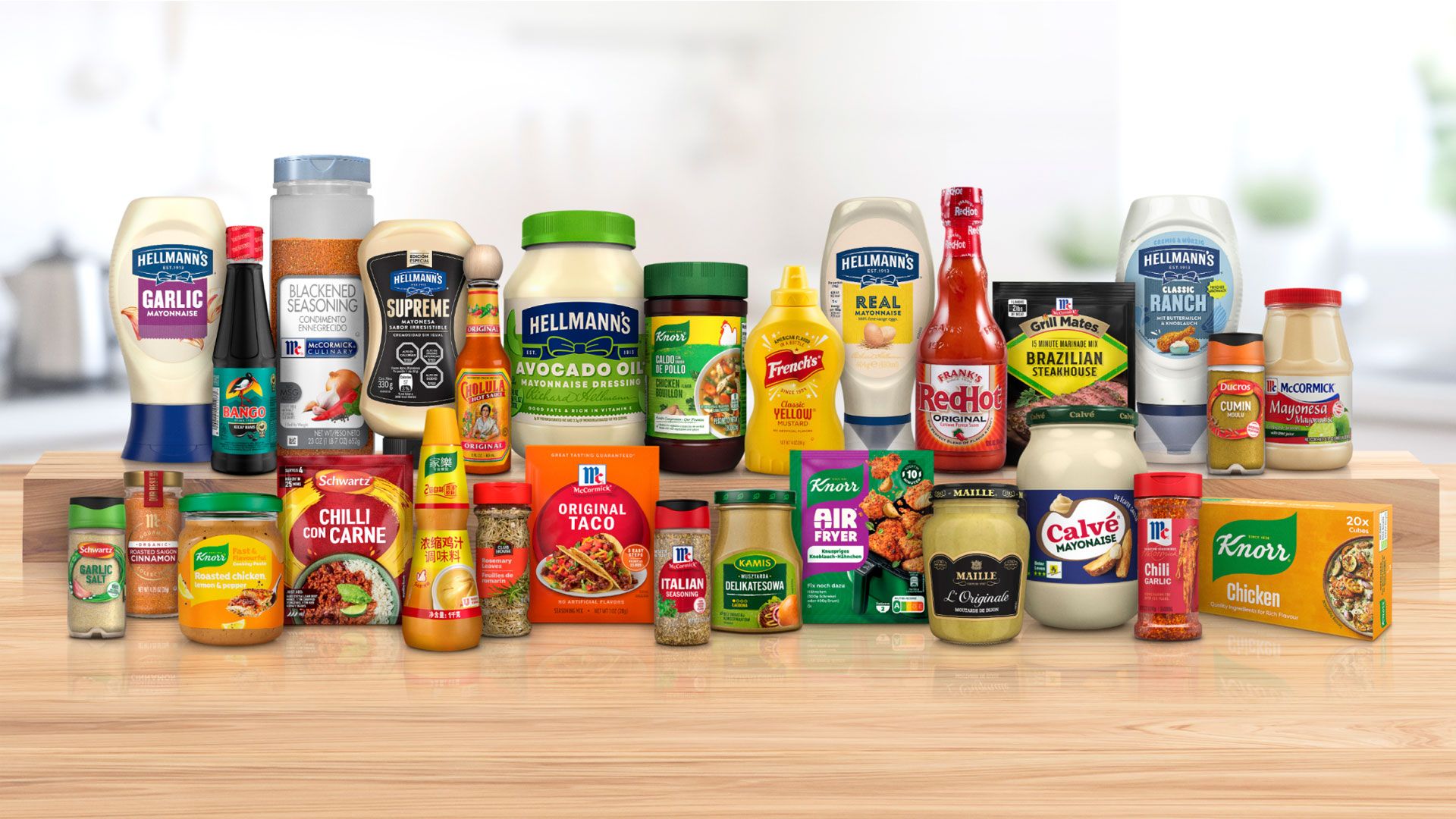 A selection of well known food and condiment brands, including Hellmann’s mayonnaise, Frank’s RedHot, Cholula Hot Sauce, Knorr seasonings, French’s mustard, spices and condiments, arranged side by side on a wooden table.