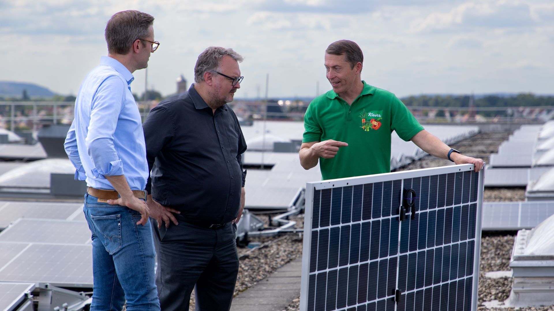 Three men standing on a rooftop covered with solar panels. One man is holding a detached solar panel.