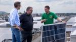 Three men standing on a rooftop covered with solar panels. One man is holding a detached solar panel.