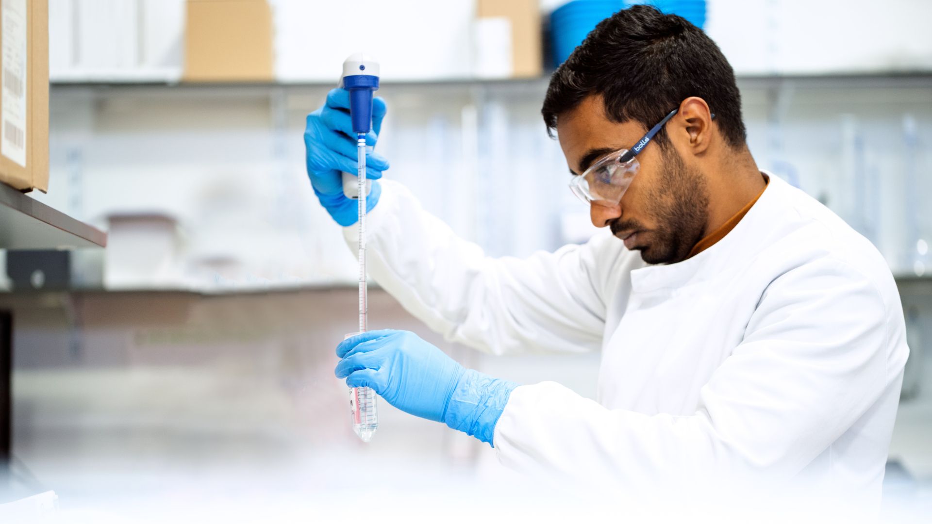 A person in a science lab wearing gloves and eye protection adding liquid to a test tube.