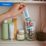 A hand reaches out to pick up a Liquid I.V. Hydration Multiplier stick in a white-and-peach design labelled ‘Sugar-Free, White Peach.’ Several other sticks are stored upright in a clear glass beside various skincare bottles, jars, and tubes. The scene includes neatly arranged personal care items and reed diffusers. The image is tagged ‘Category’.