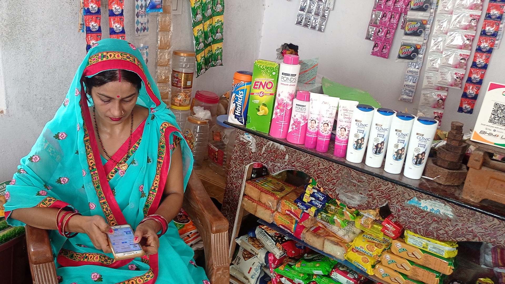 Indian woman store owner using mobile phone app while sitting next to shelves of consumer products.