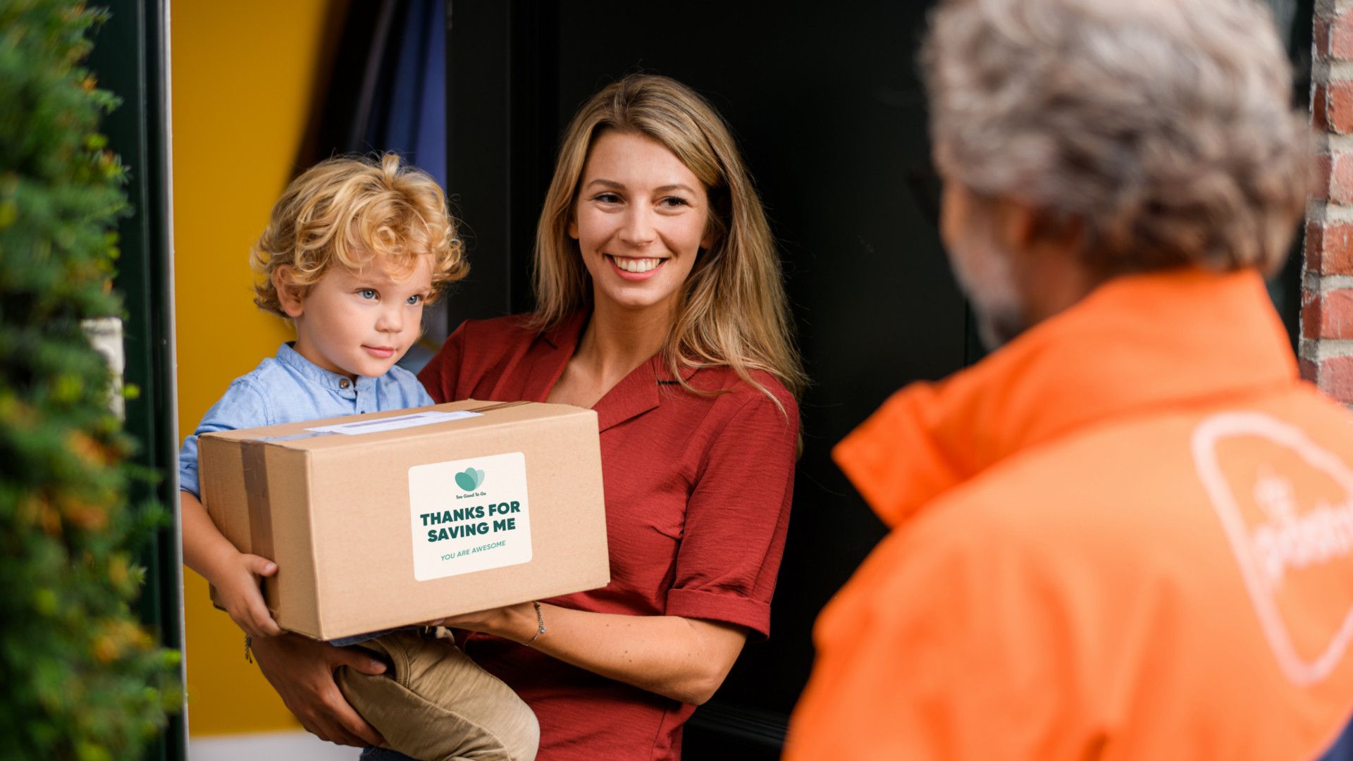 A smiling woman holds her child as she stands at her front door and accepts a parcel from Too Good To Go.