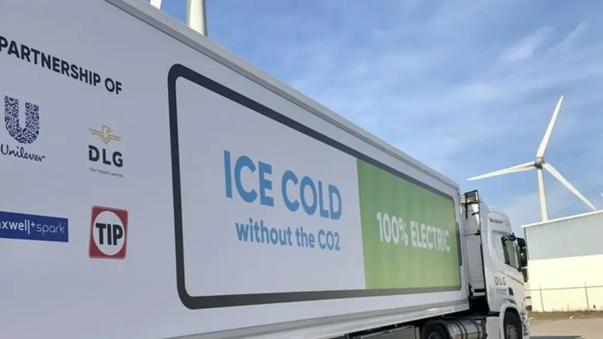 A close-up of an electric truck with a sign reading 'ICE COLD without the CO2' and '100% ELECTRIC' on its side. Logos of partnering companies, including Unilever, DLG, Maxwell + Spark, and TIP, are displayed. A wind turbine and industrial buildings are visible in the background.