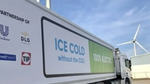 A close-up of an electric truck with a sign reading 'ICE COLD without the CO2' and '100% ELECTRIC' on its side. Logos of partnering companies, including Unilever, DLG, Maxwell + Spark, and TIP, are displayed. A wind turbine and industrial buildings are visible in the background.