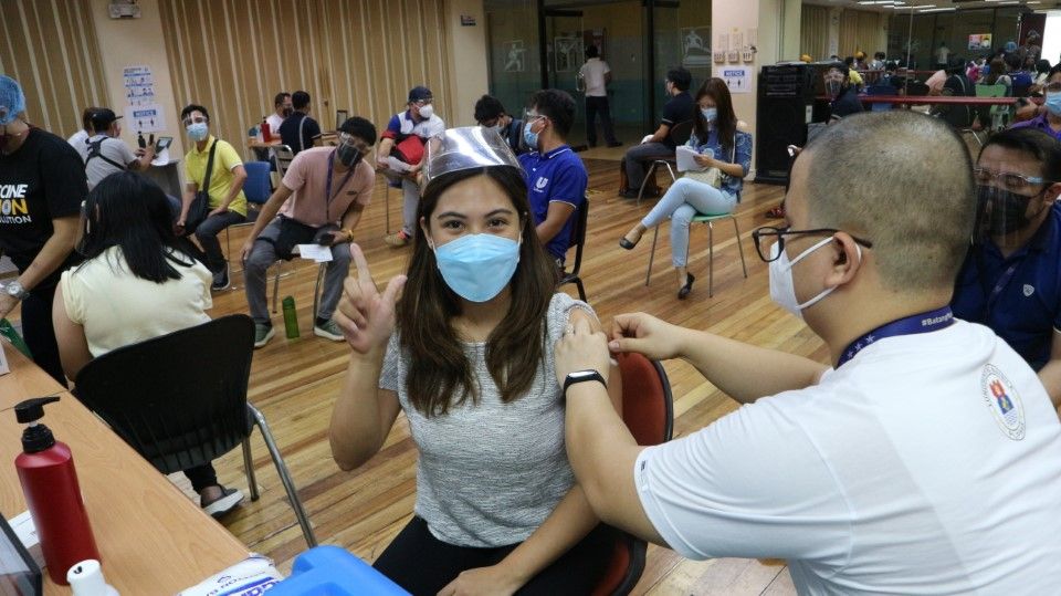 Woman being vaccinated in vacc centre, with others waiting in the background