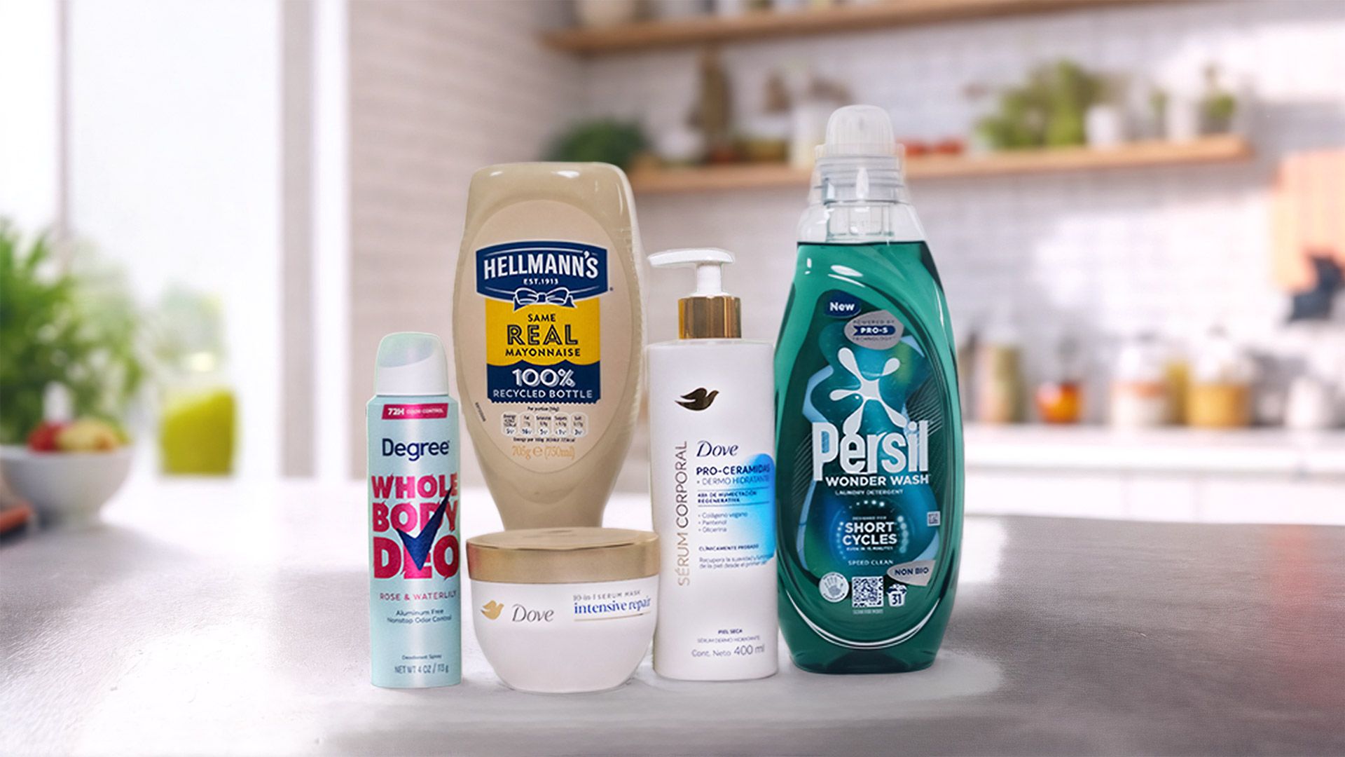 Five household and personal care products displayed on a kitchen counter: a bottle of Hellmann’s Real Mayonnaise, Degree Whole Body Deodorant spray, Dove Intensive Repair cream jar, Dove Pro-Ceramide body serum with pump, and Persil Wonder Wash laundry detergent in a green bottle. A blurred kitchen background with plants and utensils is visible.
