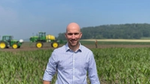 Lucas Urbano, wearing jeans and a checked shirt, stands in front of a field with two green and yellow tractors behind.