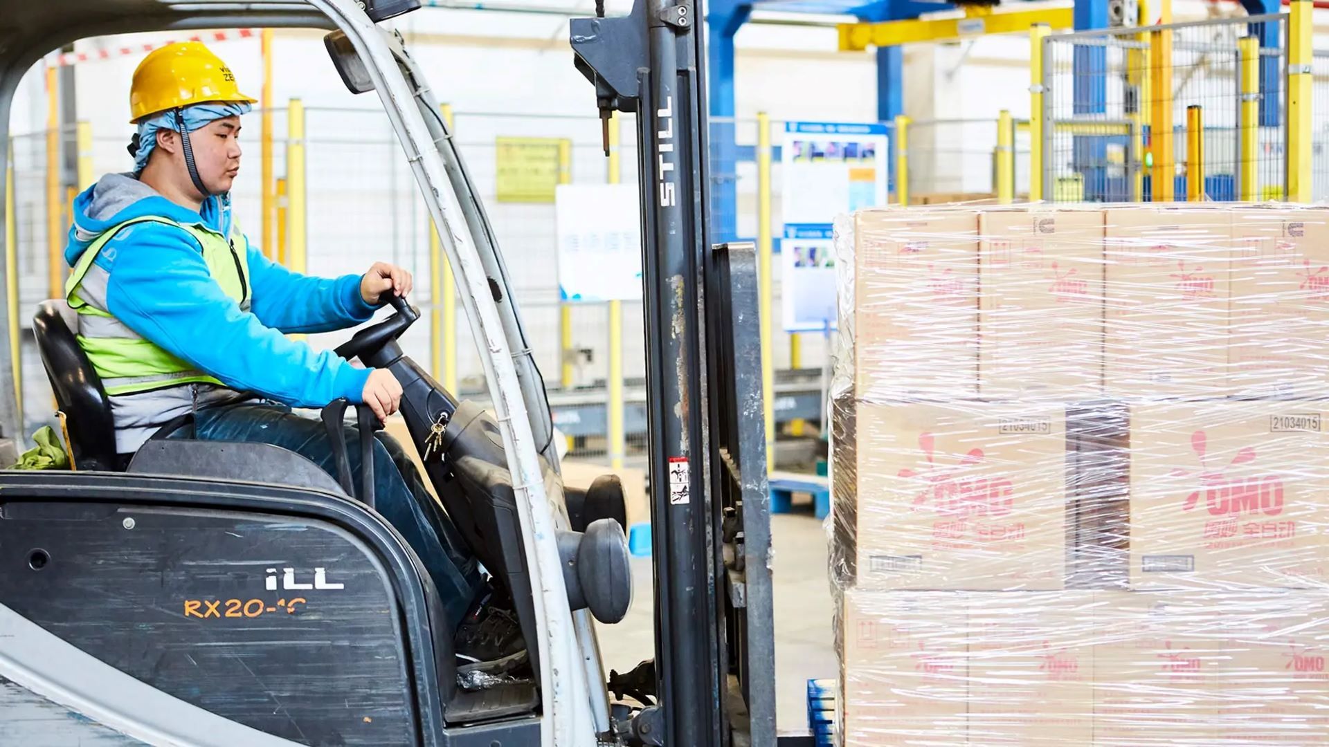 A worker operating a forklift in a warehouse, moving a pallet of neatly stacked boxes labelled ‘OMO’. The worker is wearing a yellow hard hat, a blue hoodie and a reflective safety vest, with the warehouse environment visible in the background.