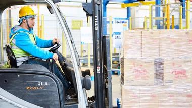 A worker operating a forklift in a warehouse, moving a pallet of neatly stacked boxes labelled ‘OMO’. The worker is wearing a yellow hard hat, a blue hoodie and a reflective safety vest, with the warehouse environment visible in the background.