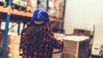 The back of the head of a young woman in a hard hat surveying a warehouse of goods
