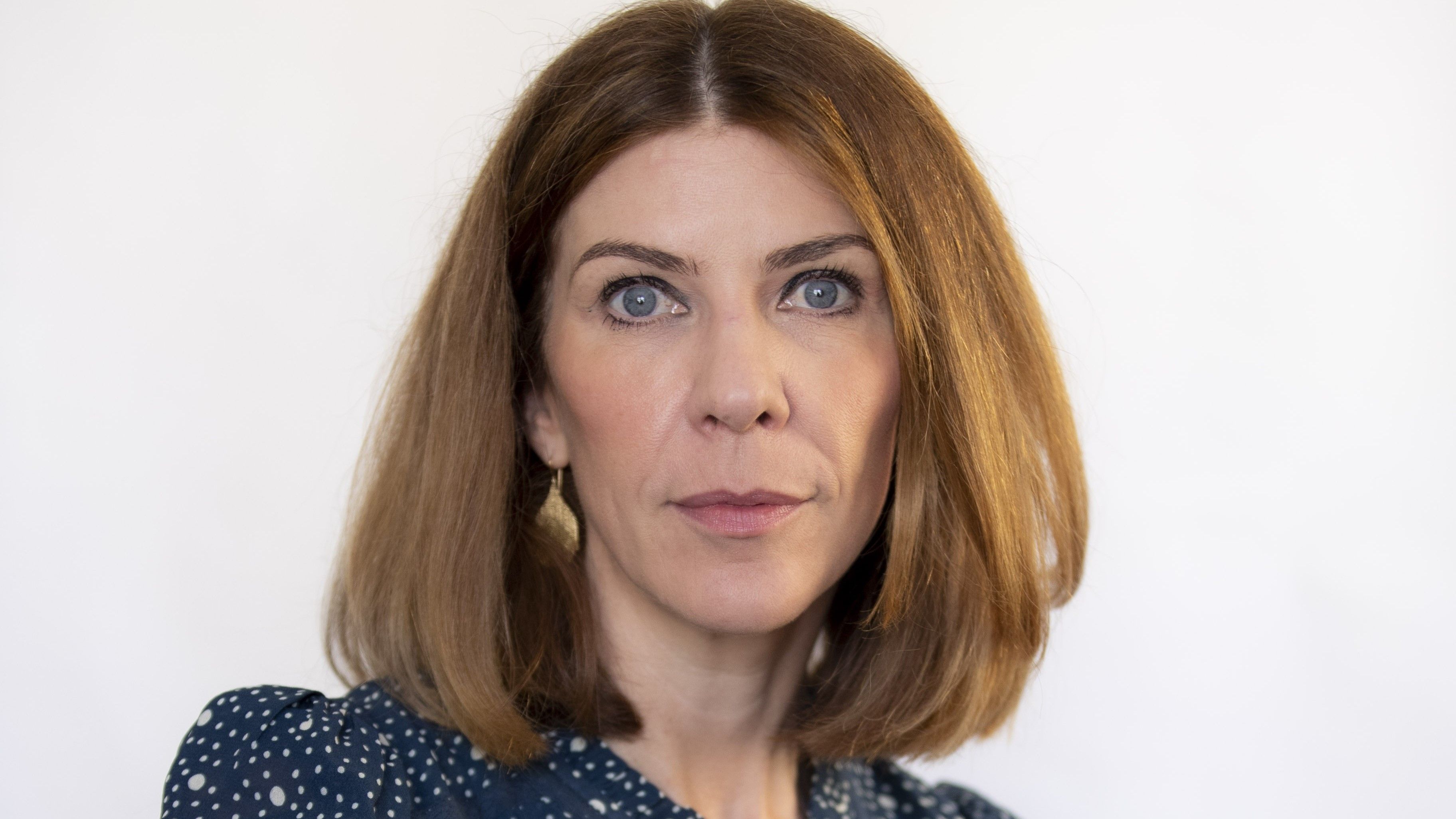 A headshot of Alison Taylor. Alison has short brown hair and blue eyes, and is wearing a navy blue shirt with white spots on. She is resting her right arm on the edge of the seat as she looks towards the camera.