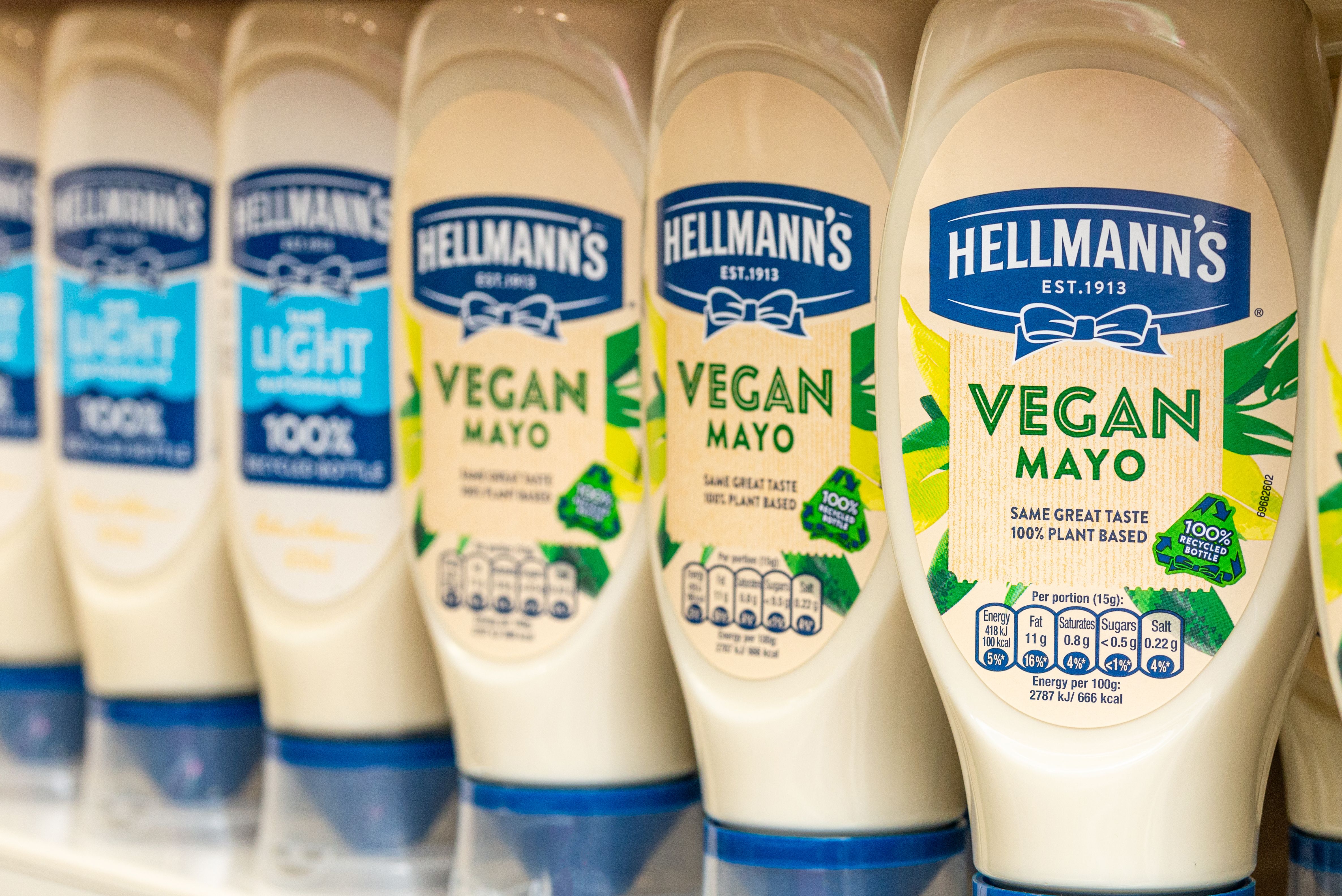 A close-up of Hellmann’s mayonnaise bottles on a shelf, featuring a mix of ‘Light’ and ‘Vegan Mayo’ varieties. The Vegan Mayo bottles are labelled with ‘Same Great Taste, 100% Plant-Based’ and ‘100% Recycled Bottle’.
