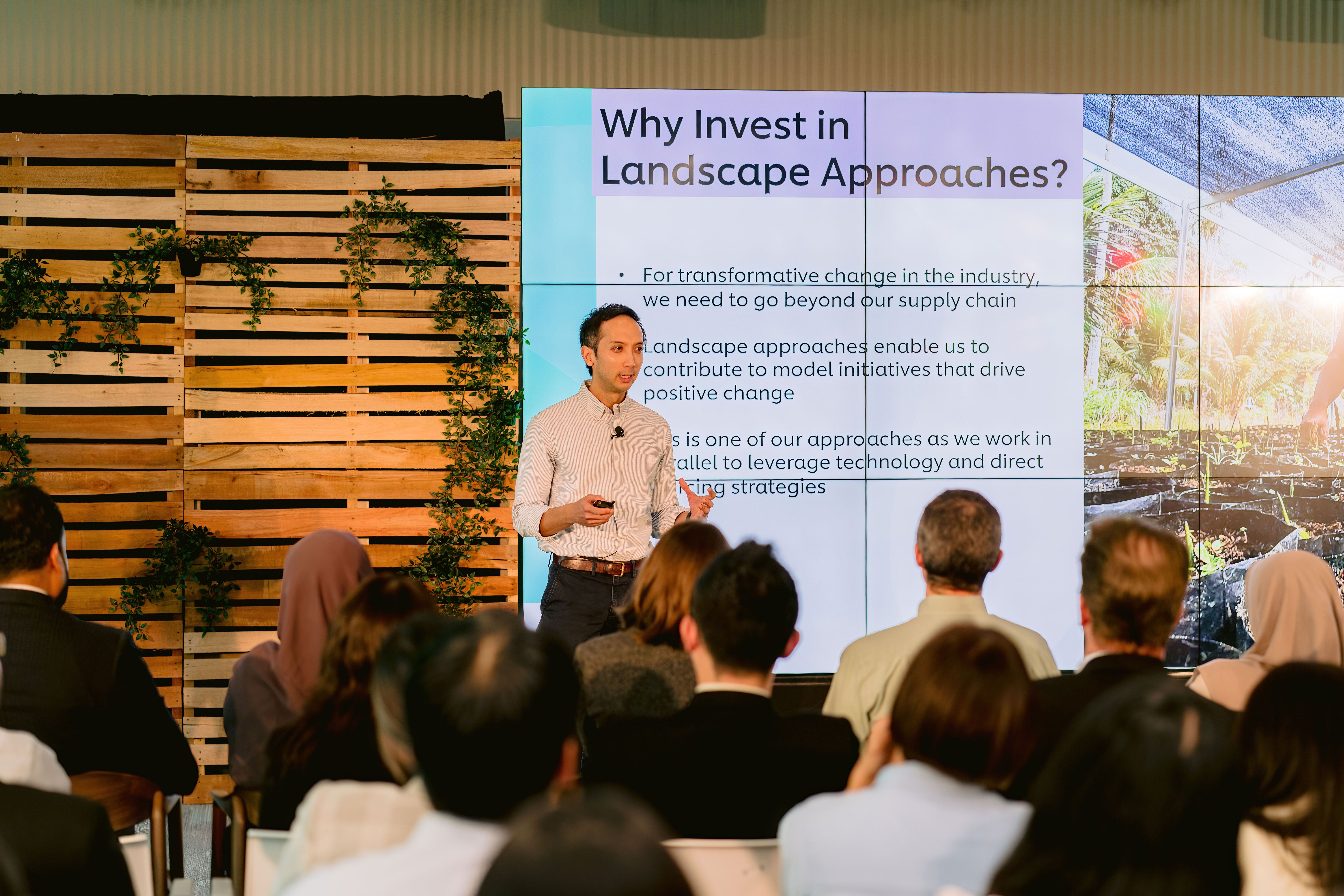 A presenter stands on a stage addressing an audience, gesturing toward a large digital screen displaying a presentation slide titled "Why Invest in Landscape Approaches?". To the left of the screen, the stage backdrop features a rustic wooden pallet wall decorated with green climbing vines.