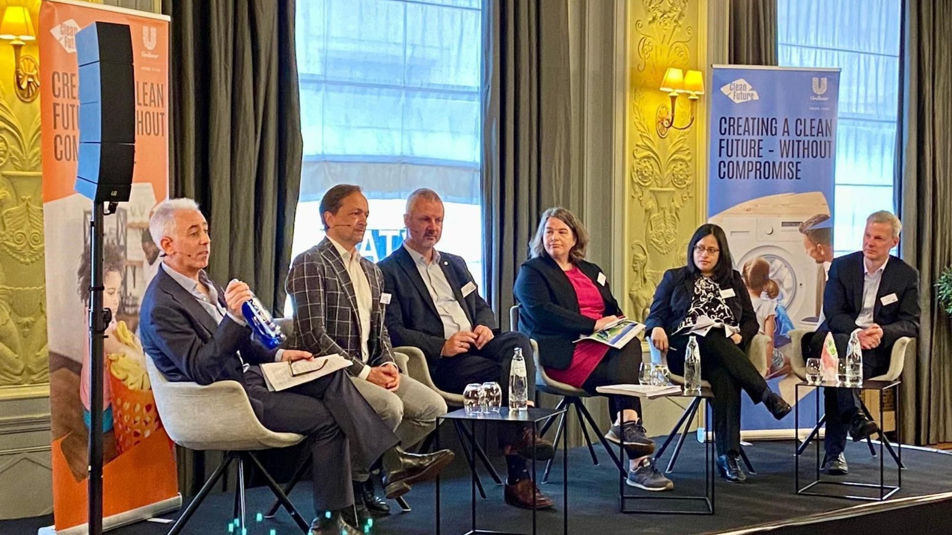 Six panellists seated on a stage at an event, with banners in the background displaying the text 'Creating a clean future – without compromise.'
