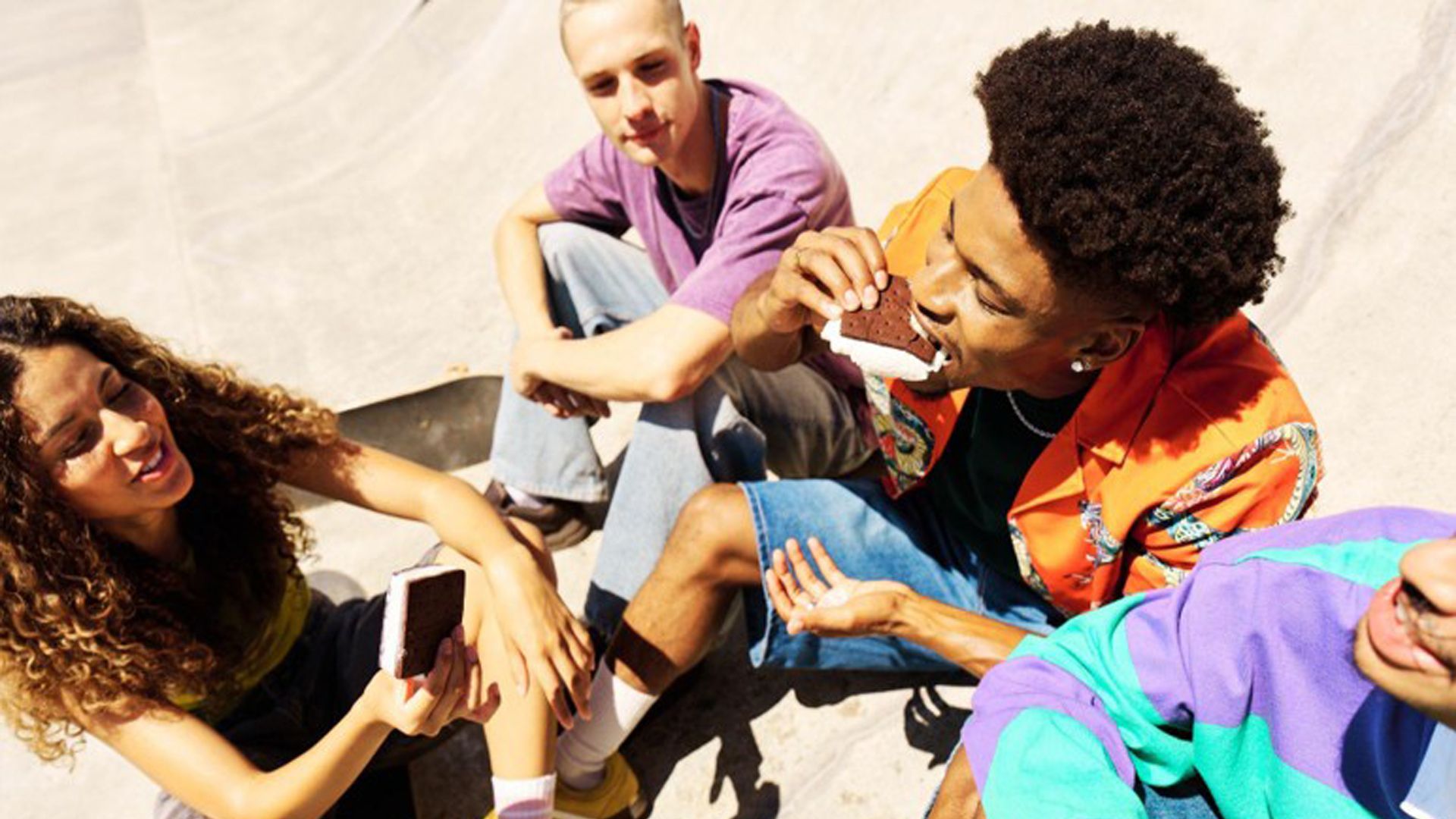 Four casually dressed individuals sitting on a concrete surface, possibly at a skate park, eating ice creams.