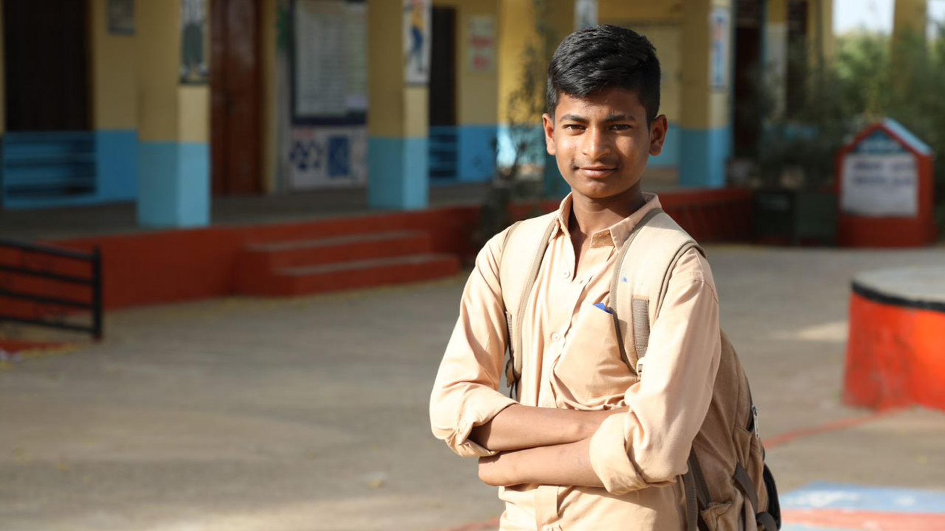 14-year-old Virendra standing with his arms folded, carrying a bag on his shoulder outside his school. 