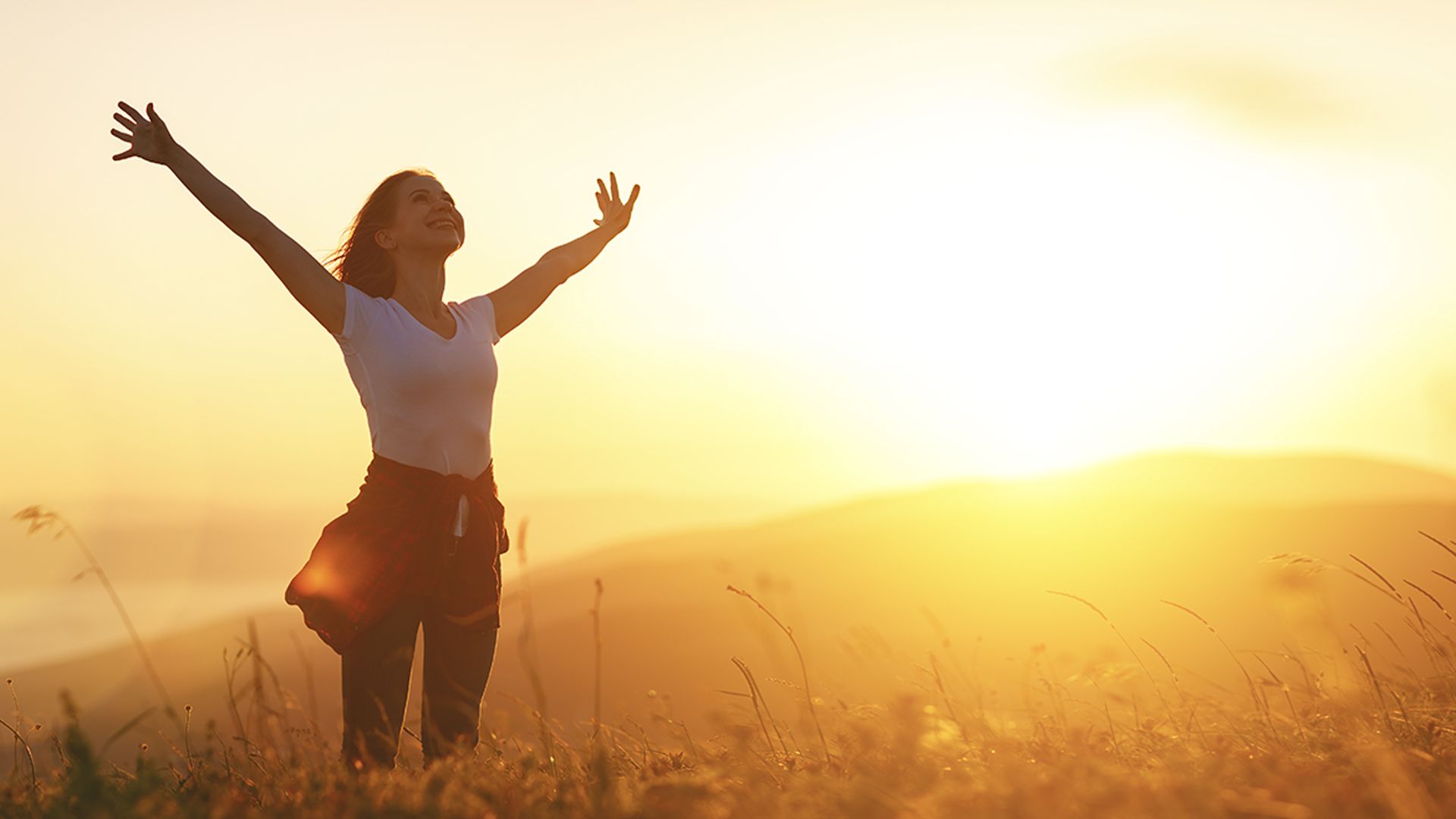 A person standing in a field with her arms raised. The sun is setting behind them casting a bright orange glow across the countryside.
