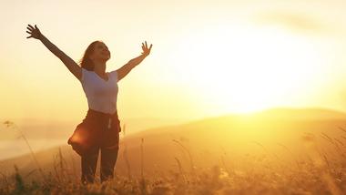 A person standing in a field with her arms raised. The sun is setting behind them casting a bright orange glow across the countryside.